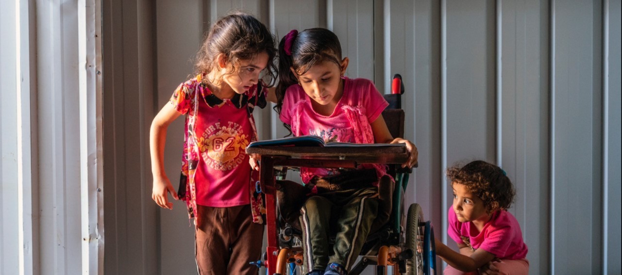A young girl sitting in her wheelchair while reading a book to a younger girl standing besides her.