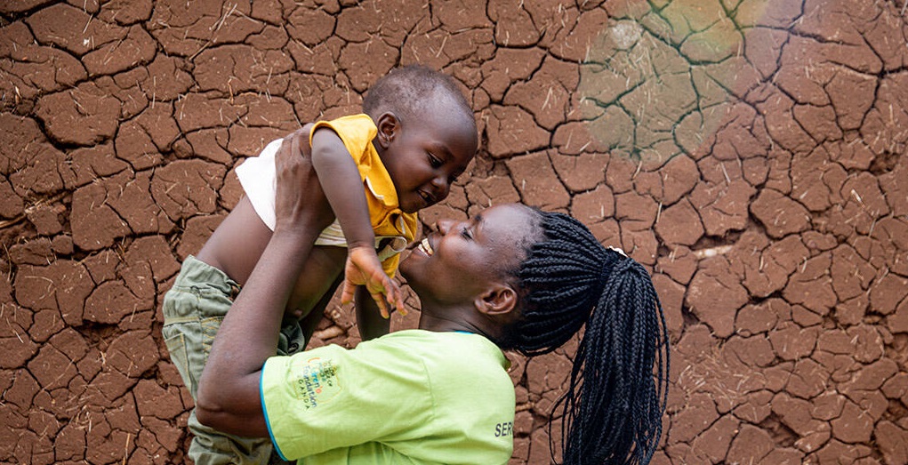 Woman with dark skin looking happy holds young child in the air