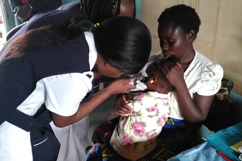 Nurses performing an ear examination on infant who sits in mothers lap, Zambia