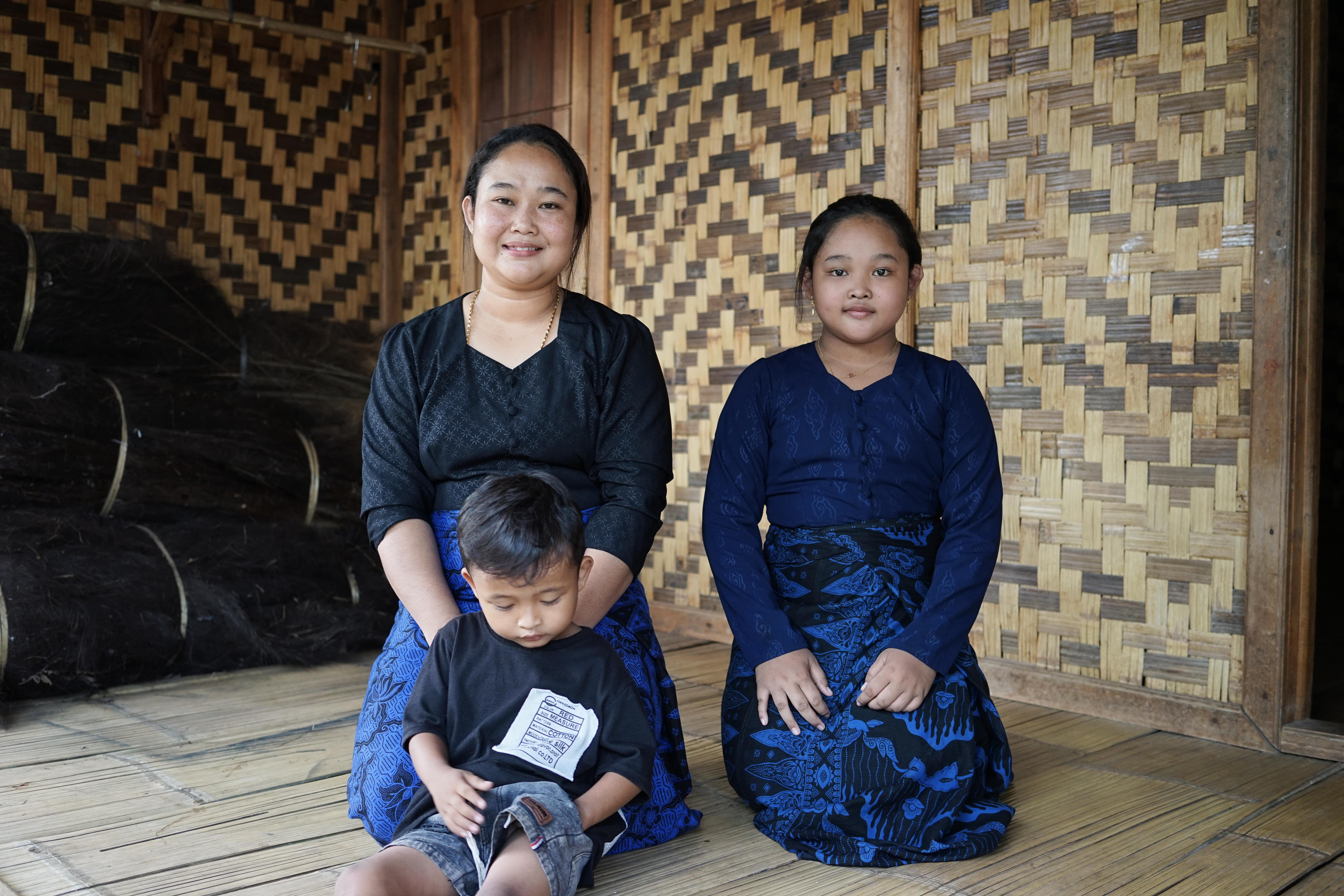 Dua perempuan dan seorang anak laki-laki duduk di lantai kayu dengan latar belakang dinding anyaman bambu.