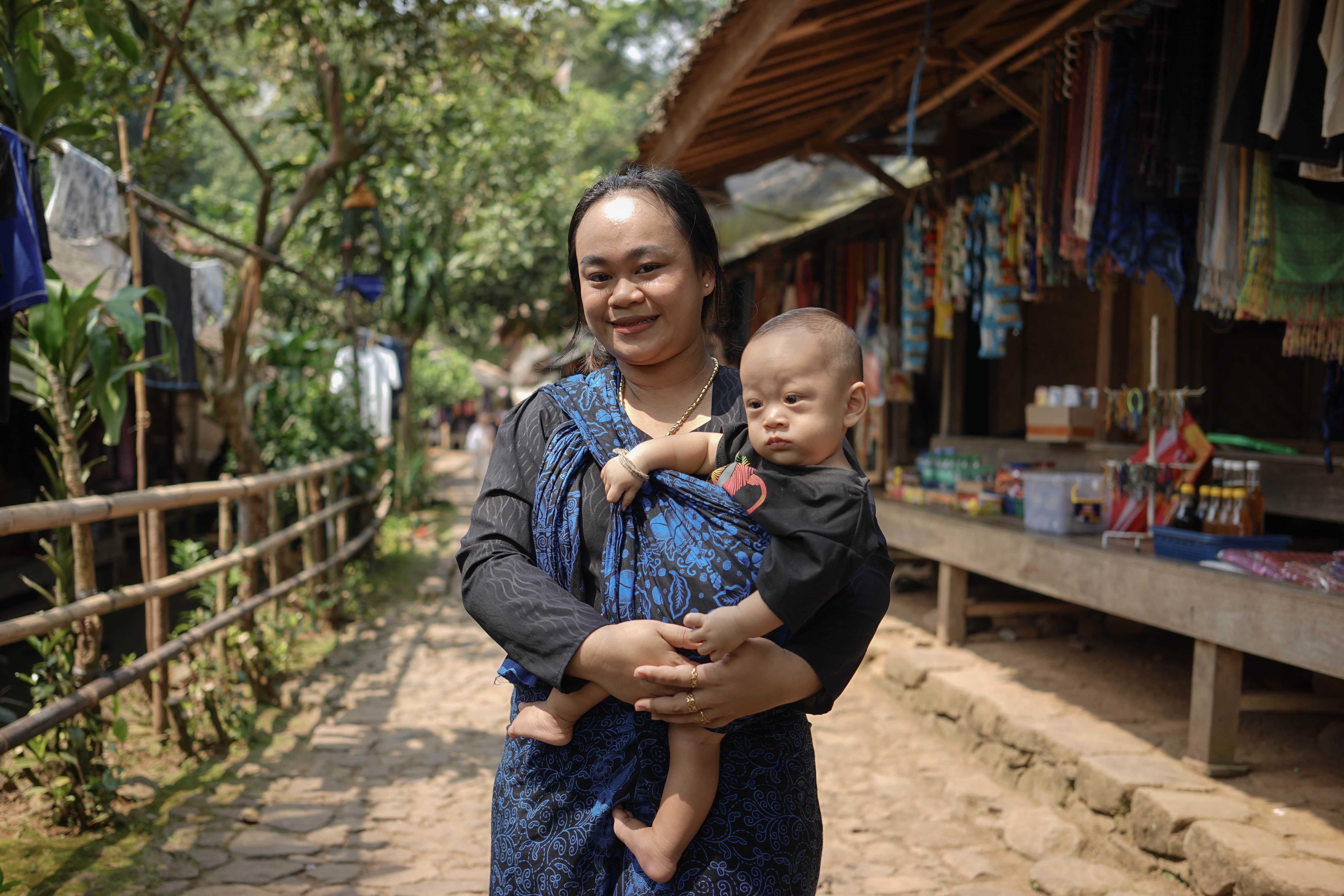 Seorang perempuan mengenakan kebaya hitam dan biru sedang menggendong bayi di depan warung tradisional.