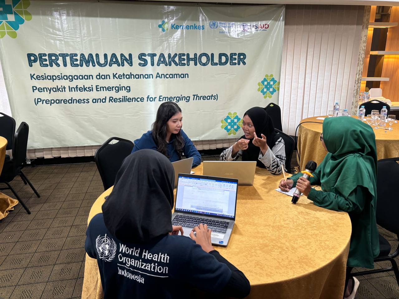 Four participants are sitting around a round table, discussing with a laptop and a microphone, taking notes